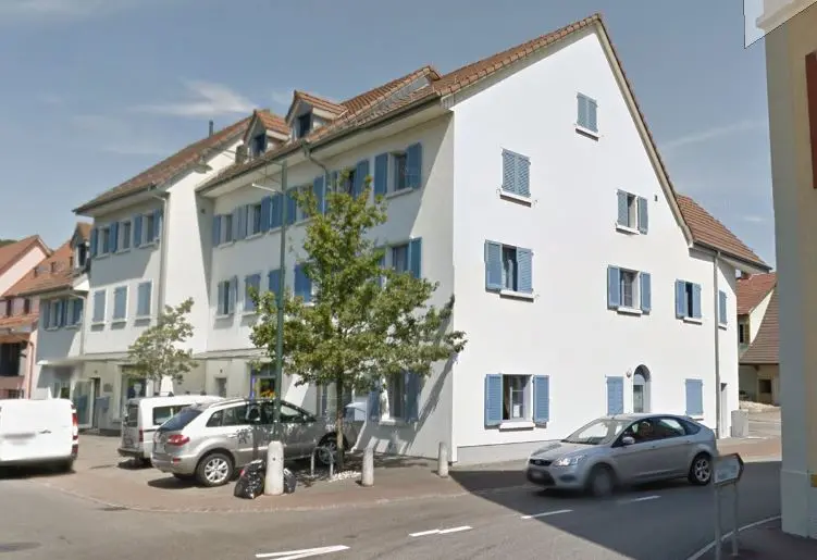 Alt-Tag: Wohn- und Geschäftshaus mit Parkplätzen und Fensterläden im Ortskern