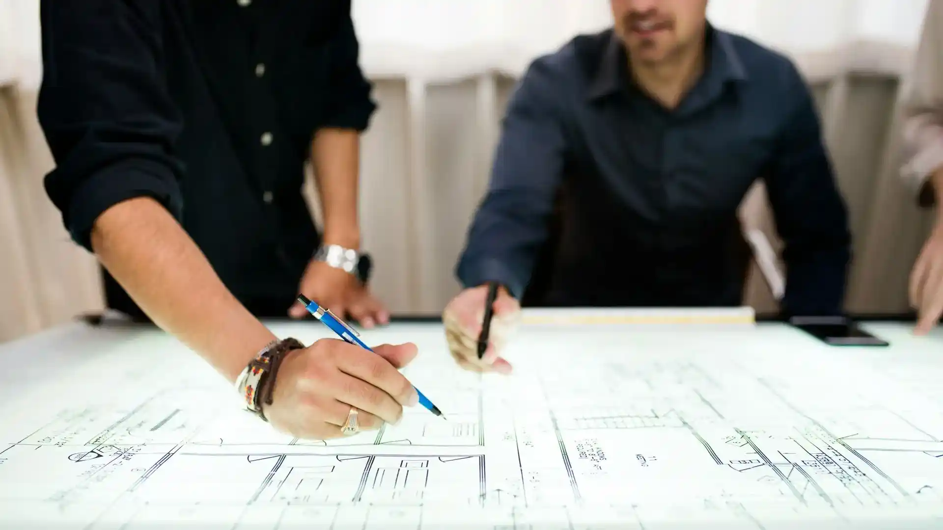 wei Personen bei der gemeinsamen Planung von Architekturzeichnungen am Tisch – eine Person mit Bleistift, die andere zeigt auf die Pläne.